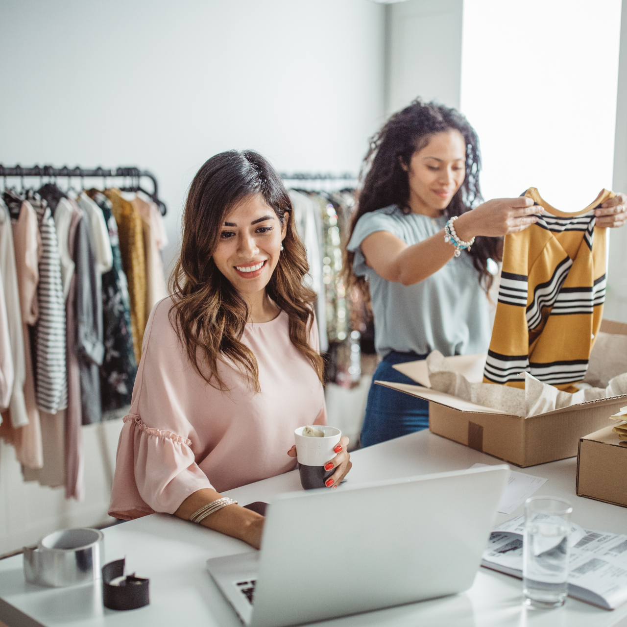 A clothing store owner is checking online orders on a laptop, while a staff member nearby is organizing clothes in the store